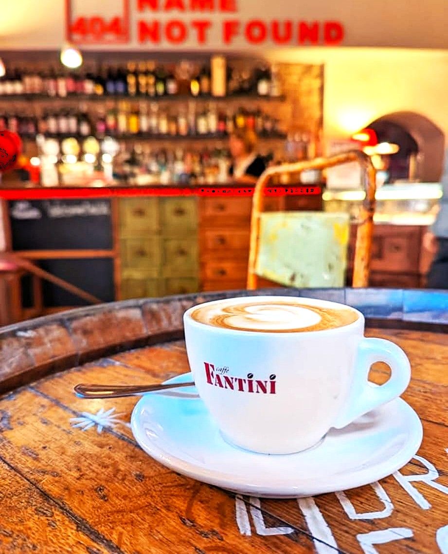 A cup of coffee with 'Fantini' written on it, placed on a wooden table with a spoon.