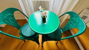 Una mesa azul verdosa con dos sillas, un florero con flores y una ventana con cortinas.