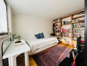 A bedroom with a bed, a small desk, a bookshelf, and a large window. - Torre Tolomei Trastevere (Rome)