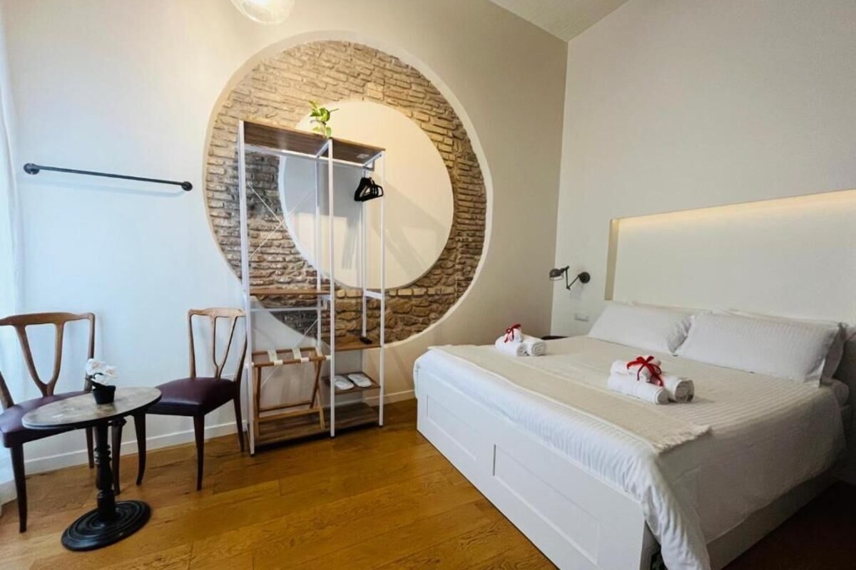 Un dormitorio con pared de piedra circular, una cama con ropa blanca, una pequeña mesa con sillas y una estantería con objetos.