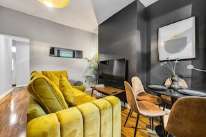 A modern living room with a yellow sofa, a dining table with chairs, a flat-screen TV, and a framed artwork on the wall. - 3 BR Home 3 min Close to MCR City Centre Free Park (Manchester)