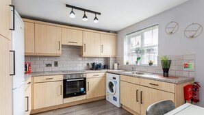 A modern kitchen with wooden cabinets, a built-in oven, a washing machine, a granite countertop, and a window with blinds. - Trafford Centre Cityscape Hideaway Free Parking (Manchester)