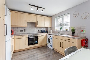 A modern kitchen with wooden cabinets, a built-in oven, a washing machine, a granite countertop, and a window with blinds.