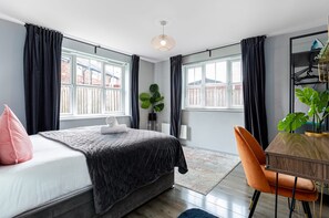 A bedroom with a bed, a desk, and a chair. There are windows with curtains, a plant, and a ceiling light.