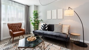 Ein modernes Wohnzimmer mit Ledersessel, dunklem Sofa, Glastisch, Stehlampe und Zimmerpflanze.