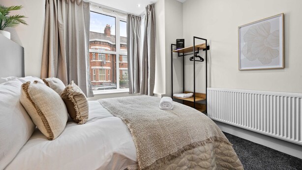 A hotel room with a bed, a radiator, a window with curtains, a coat rack, and a framed picture on the wall. - Sleeps 14 The Old Trafford Zen by Genesis Living (Manchester)