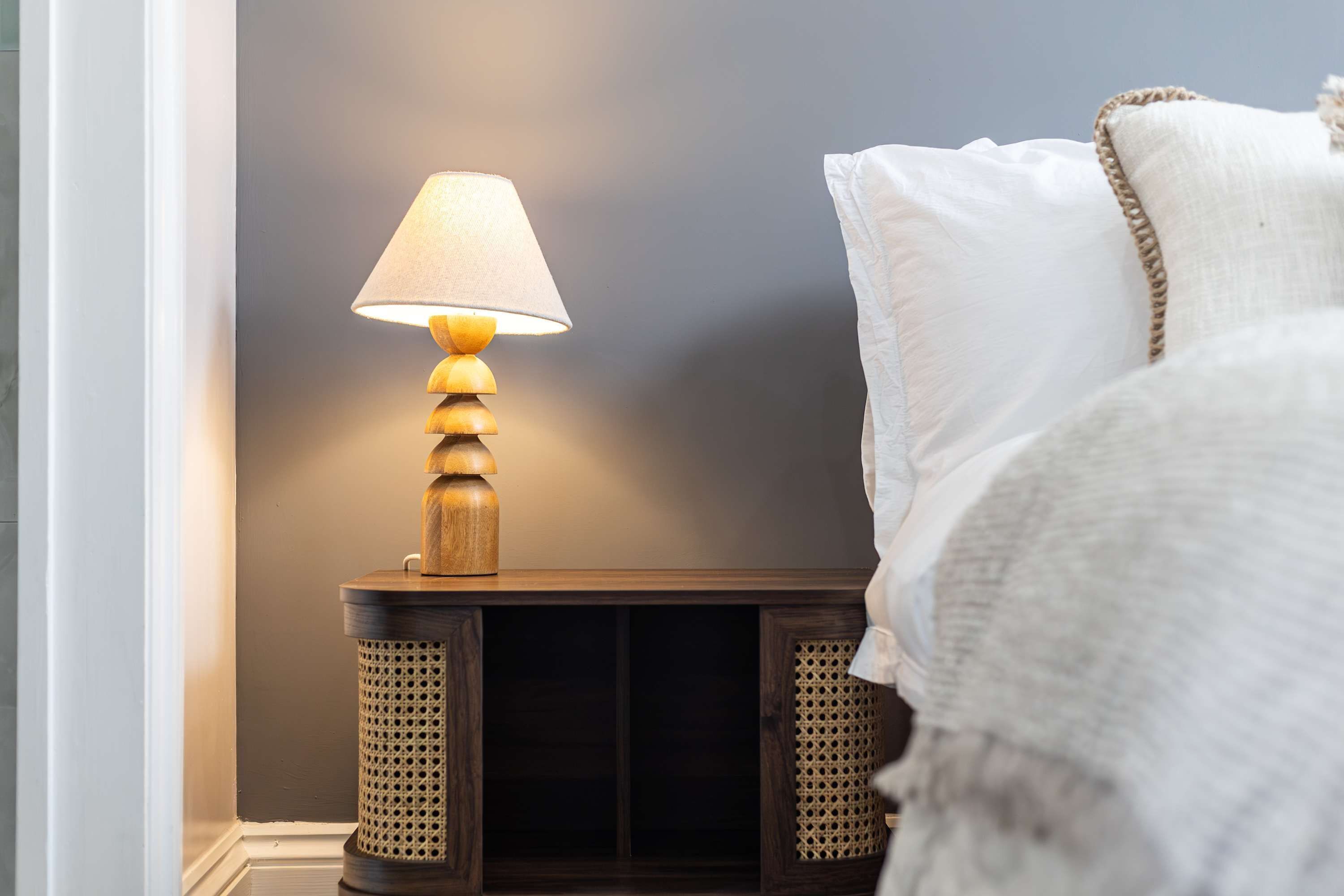 Une table de chevet avec une lampe, un lit avec du linge de lit blanc et un mur foncé.