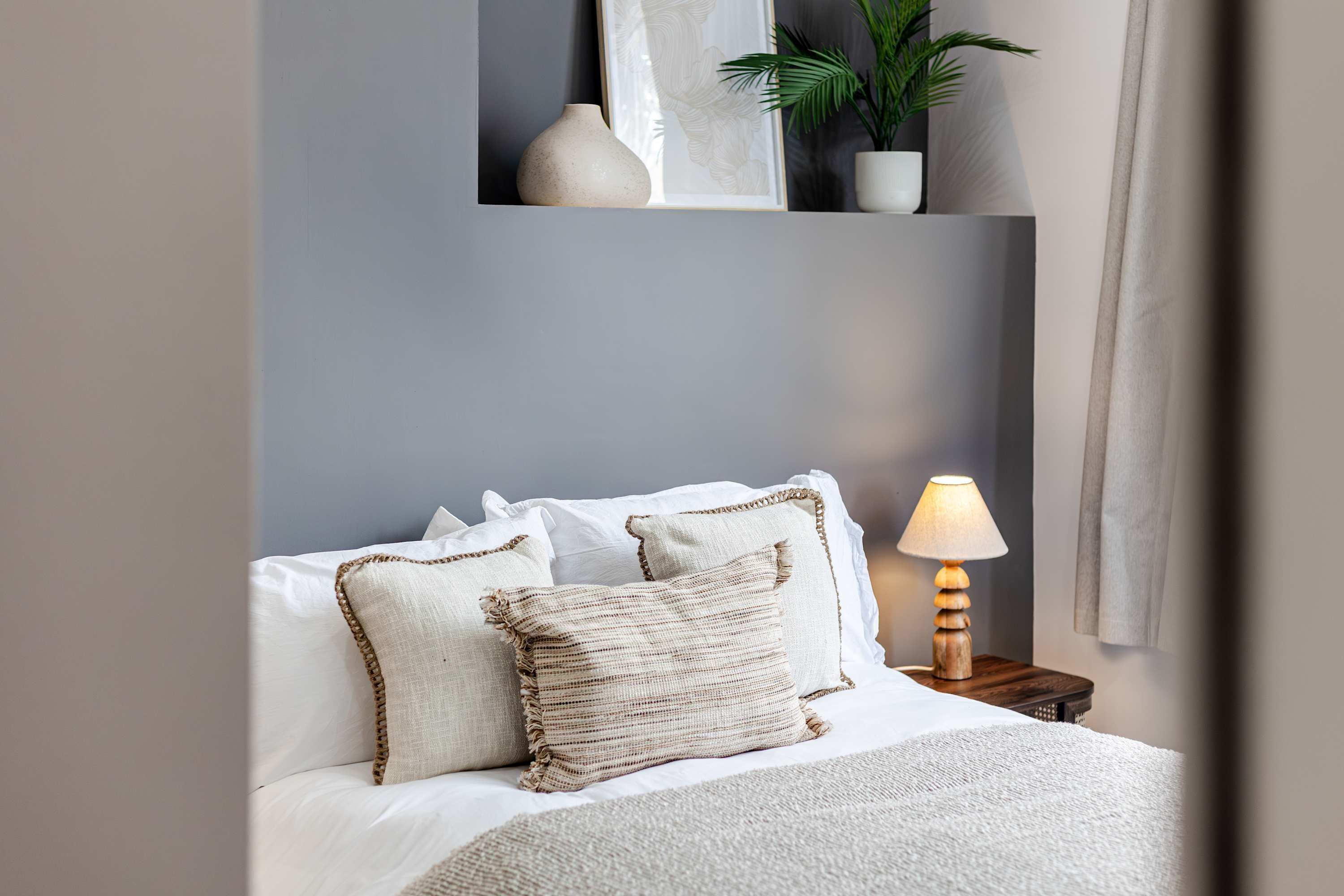 Een netjes opgemaakt bed met witte lakens en twee decoratieve kussens, een nachtkastje met een lamp, en een wandplank met een plant en een vaas.