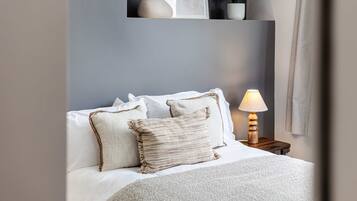 Een netjes opgemaakt bed met witte lakens en twee decoratieve kussens, een nachtkastje met een lamp, en een wandplank met een plant en een vaas.