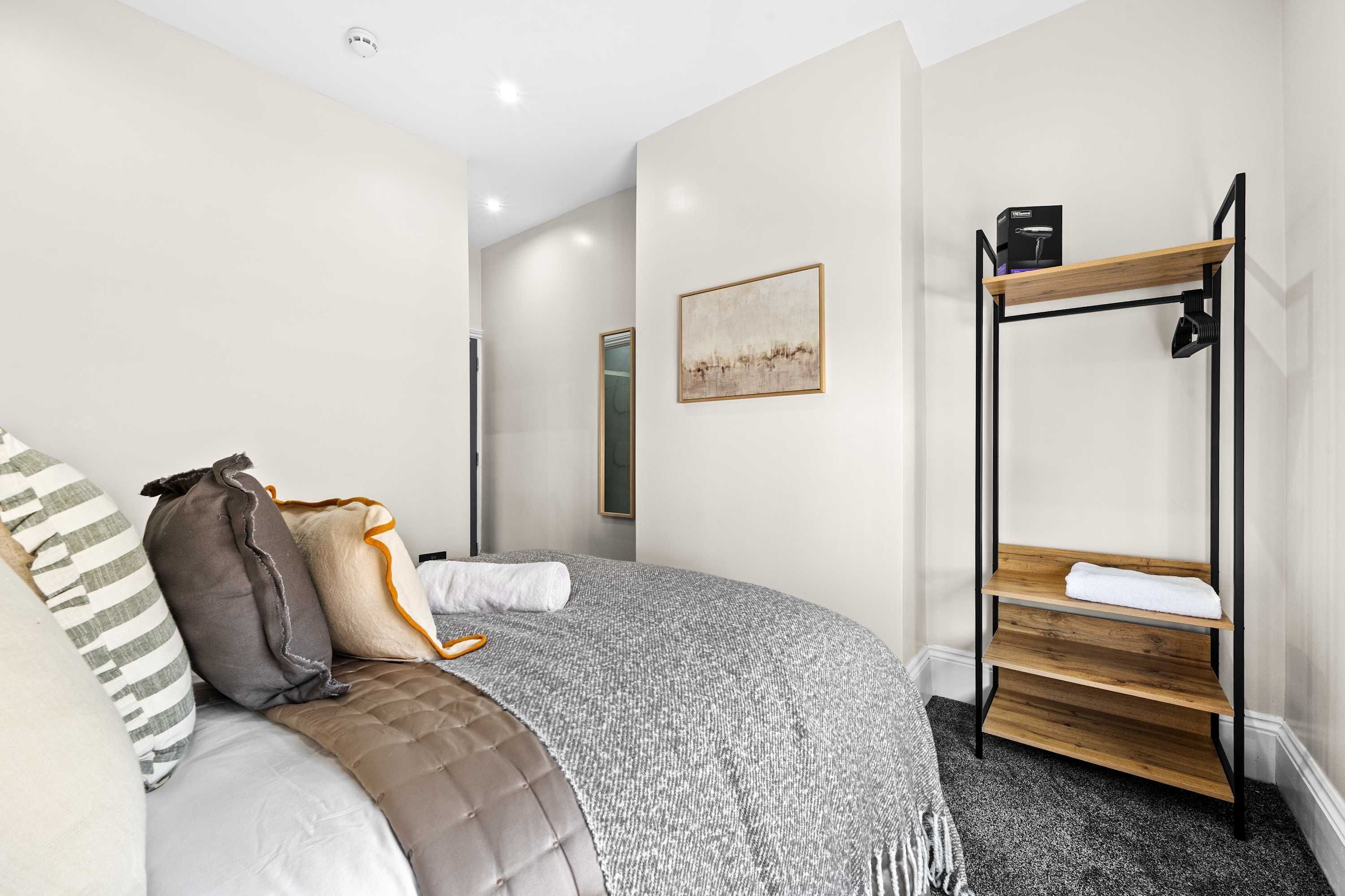 A bedroom with a bed, pillows, a blanket, a wooden shelf, and a painting on the wall.