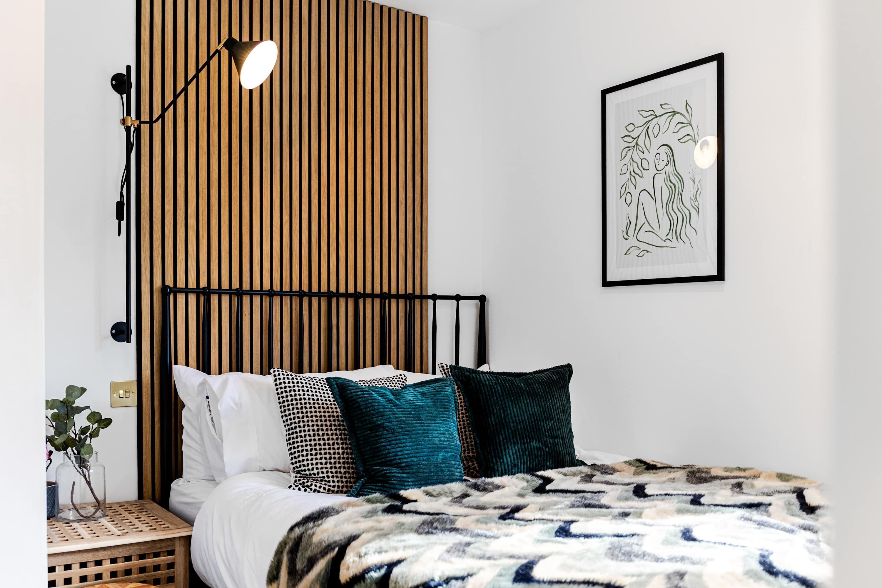 Un dormitorio con cama, almohadas, mesita de noche con lámpara, una obra de arte enmarcada en la pared y panel de pared de listones de madera.