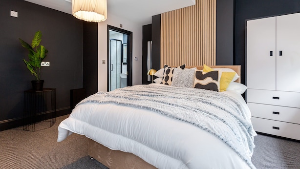 A modern bedroom with a large bed, a bedside table, a potted plant, a wardrobe, and a door leading to another room.