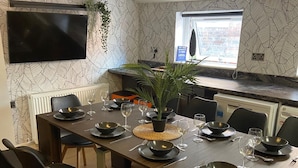 A modern dining area with a long table set for six, a kitchen with a window, and a mounted TV.