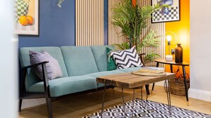 A modern living room with a teal sofa, patterned cushions, a wooden coffee table, a patterned rug, a potted plant, and artwork on the walls.