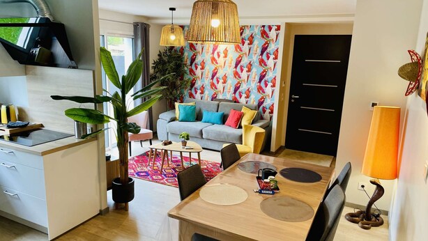 A modern kitchen with a built-in stove, a dining area with a wooden table and chairs, a grey sofa with colorful cushions, and a vibrant wall with a bird pattern.