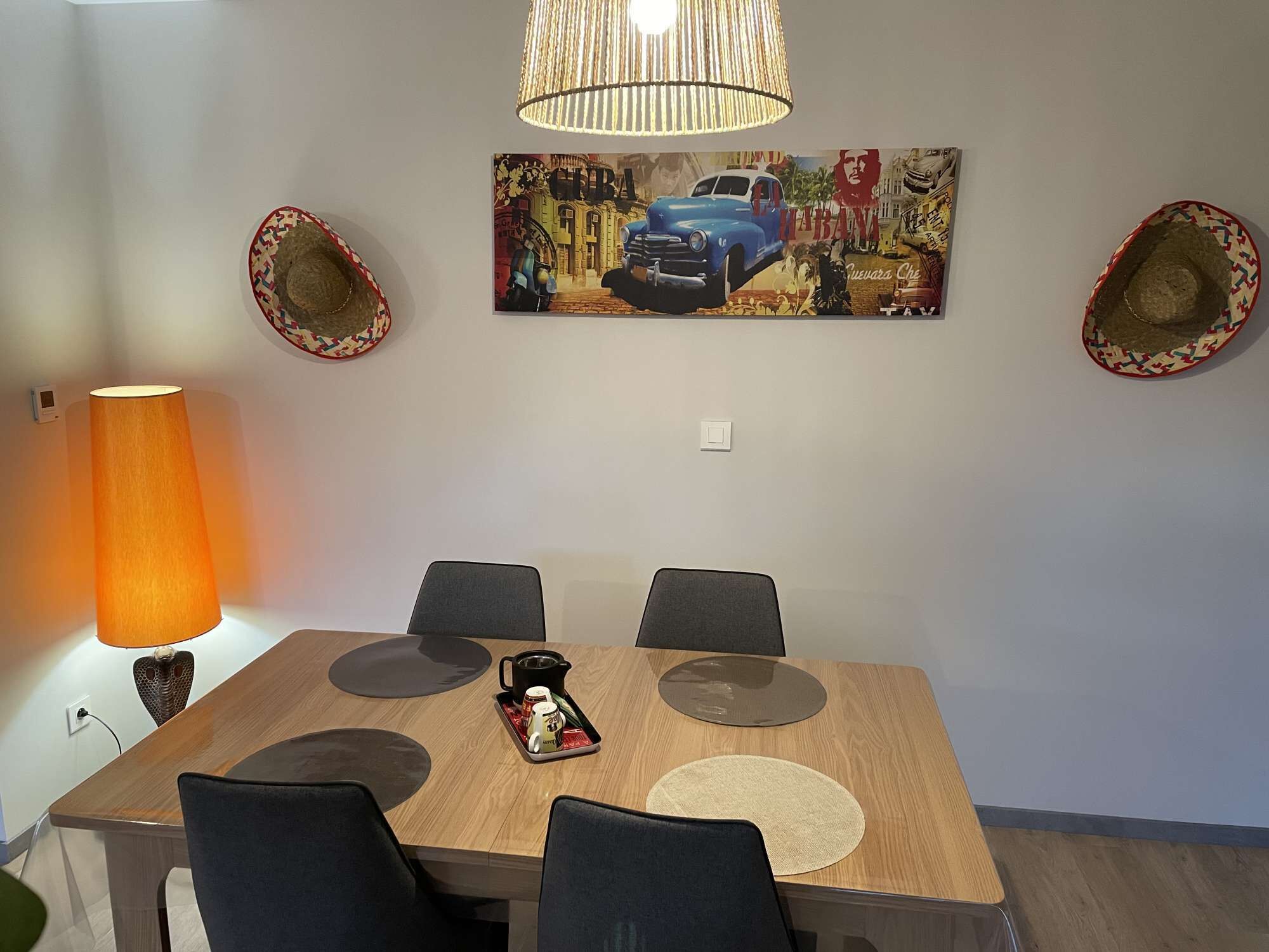 A dining area with a wooden table, four chairs, a painting of a car and cityscape, two hats on the wall, and a lamp.