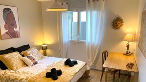 A bedroom with a bed, bedside lamps, a window with curtains, a small table with a lamp, and a wall decoration.