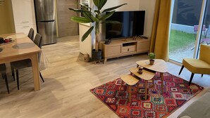 A modern living room with a TV, dining table, chairs, a coffee table, a large plant, and a colorful rug.