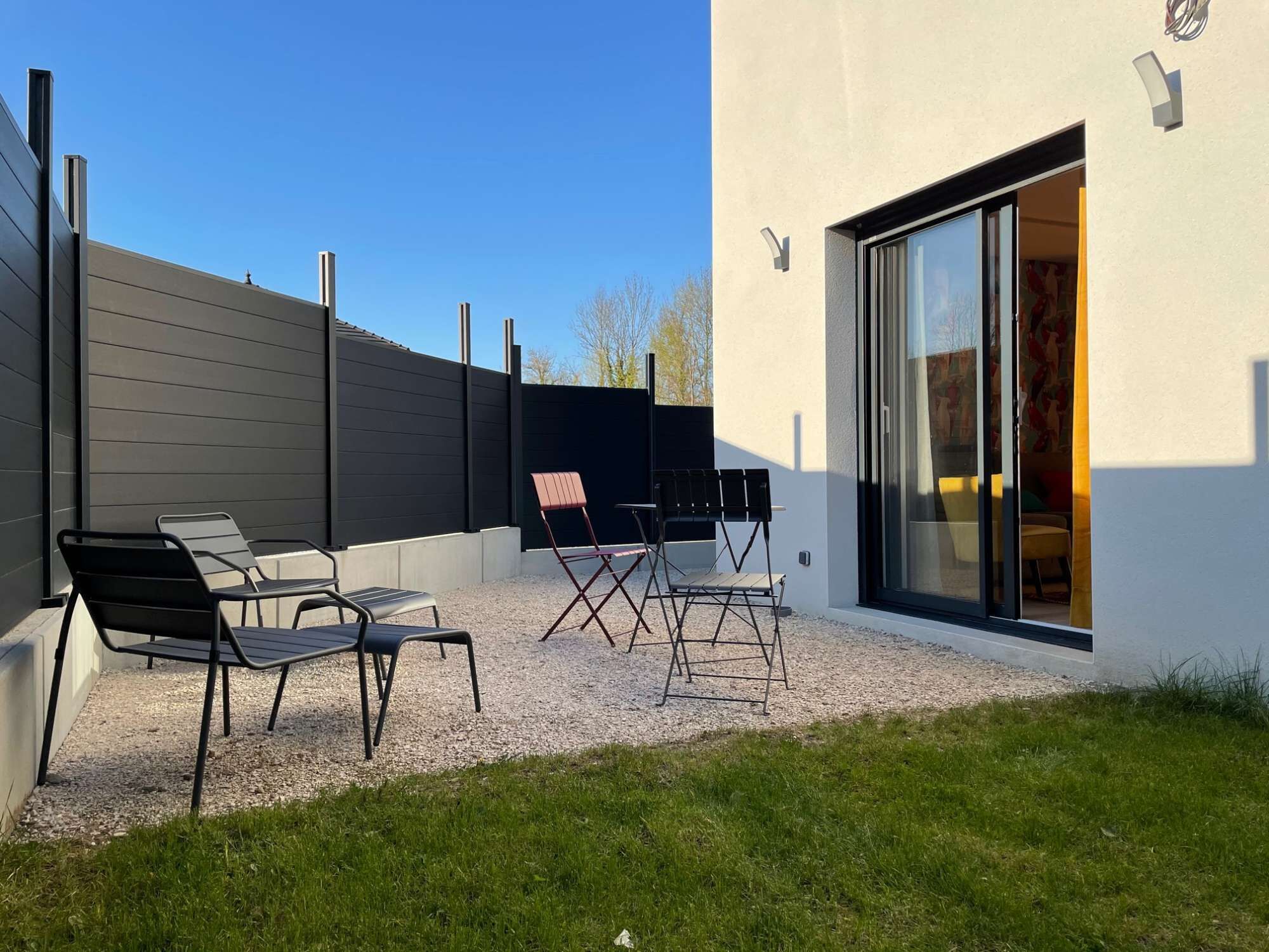 Une terrasse extérieure moderne, avec des chaises et une table en métal, un sol en gravier et un bâtiment doté d’une grande porte-fenêtre coulissante.