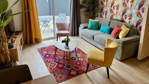 A living room with a grey sofa, colorful cushions, a patterned rug, and a large window with curtains.