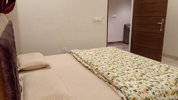 Uma cama de solteiro com colcha floral e duas almofadas, uma porta de madeira e um radiador ao fundo.