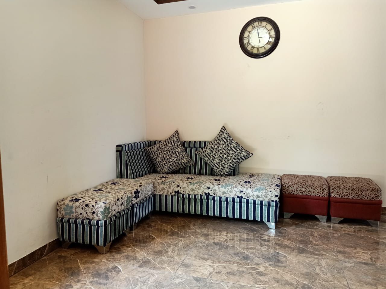 A corner room with a sofa, ottomans, a clock, and a tiled floor.