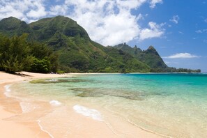 A beach with clear turquoise water, a sandy shore, and lush green mountains in the background.