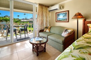 A room with a sliding glass door leading to a balcony, a sofa with a tropical print, a small table with a book, and a lamp.