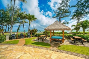 Eine Terrasse direkt am Strand mit einem reetgedeckten Pavillon, Picknicktischen und einem gepflasterten Weg.