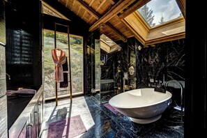 A modern bathroom with a large bathtub, a glass shower enclosure, and a view of the forest through large windows.