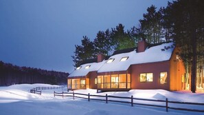 Una casa moderna de varios pisos de madera, con grandes ventanales y un tejado cubierto de nieve, rodeada de un cercado de madera y árboles.