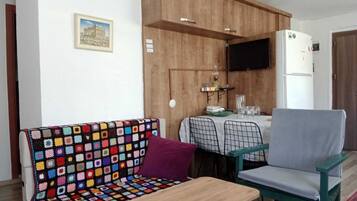 Zona tranquil·la amb un sofà de gots de colors, una taula de fusta petita i una cadira verda. Es veu una taula de menjador amb caires al fons, i un televisor fixat a la paret.