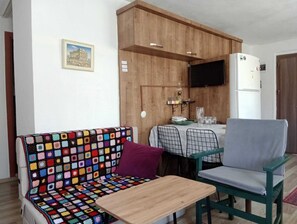 A cozy living area with a colorful crochet sofa, a small wooden table, and a green chair. A dining table with chairs is visible in the background, and a TV is mounted on the wall.