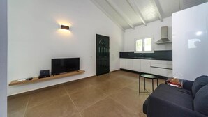 A modern living room with a flat-screen TV, a sofa, a small table, and a kitchen area with cabinets and a window. - Mary's Country House (Lefkimmi)