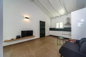 A modern living room with a flat-screen TV, a sofa, a small table, and a kitchen area with cabinets and a window. - Mary's Country House (Lefkimmi)