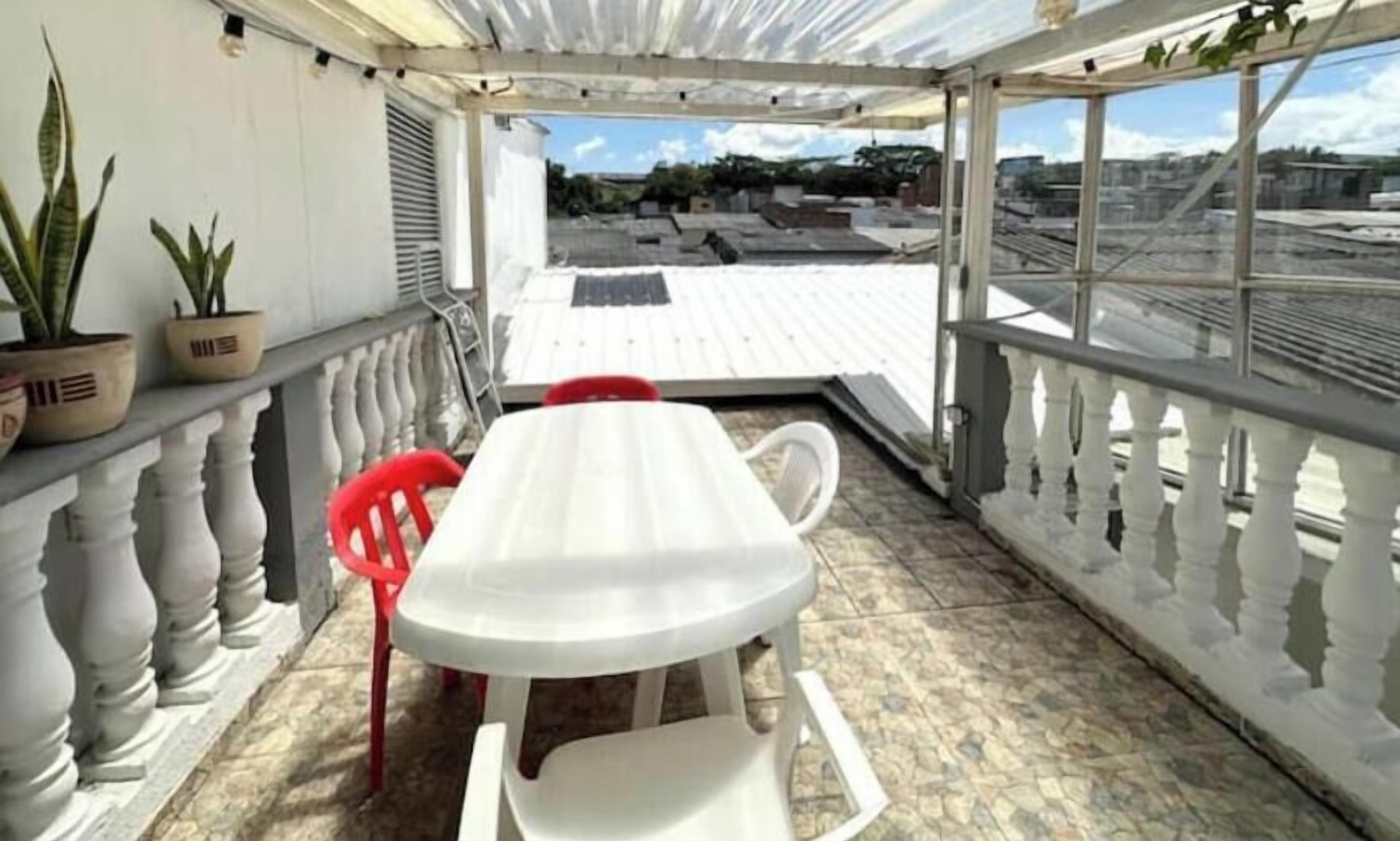 Un balcón techado con una mesa blanca, una silla roja, plantas en macetas y vista a la ciudad.