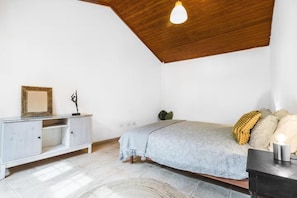 A bedroom with a sloped ceiling, a bed with patterned bedding, a small cabinet, a plant, and a light fixture.