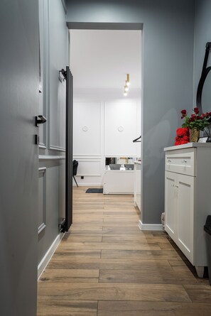 Un pasillo con piso de madera, paredes blancas, un radiador negro y un mueble blanco con un florero de flores rojas.