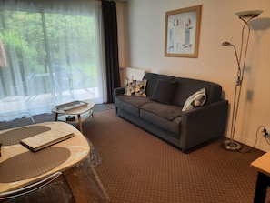 A living room with a sofa, a table with a menu, a floor lamp, and a window with a view of greenery. - Apartment AB du parc of 51 M2 sunny - with beautiful terrace of 15M2 (Neufchâtel-Hardelot)