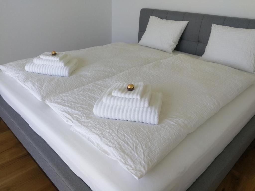 Un lit double avec du linge de lit blanc et deux serviettes, une tête de lit rembourrée de couleur grise et un mur sobre.