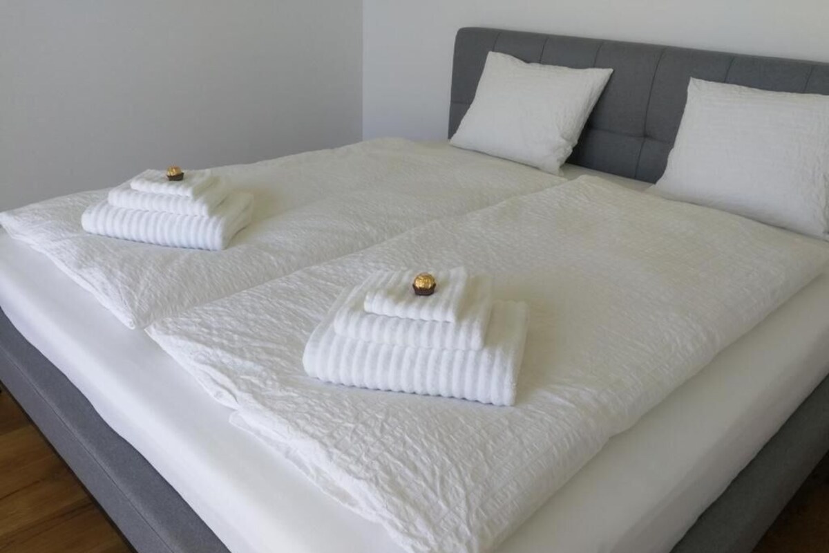 A double bed with white linens and two towels, a grey upholstered headboard, and a plain wall.