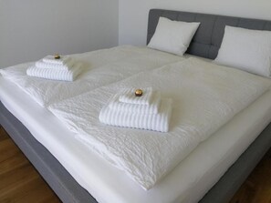 Un lit double avec du linge de lit blanc et deux serviettes, une tête de lit rembourrée de couleur grise et un mur sobre.