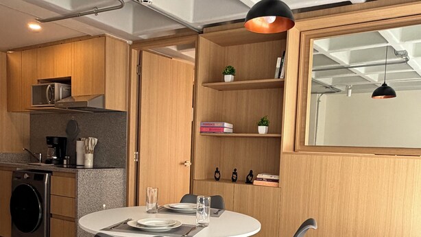 Una cucina compatta con elettrodomestici moderni, un'area pranzo con un tavolo rotondo e sedie, e una libreria a incasso con libri e oggetti decorativi.