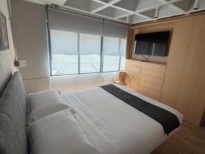 Ein Schlafzimmer mit einem Bett, einem Sessel, einem an der Wand montierten Fernseher und einem Fenster mit Jalousien.