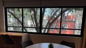 Ein Zimmer mit großem Fenster, Schreibtisch, Stuhl und rundem Tisch mit Blumentopf.