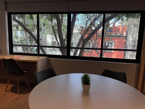 Ein Zimmer mit großem Fenster, Schreibtisch, Stuhl und rundem Tisch mit Blumentopf.