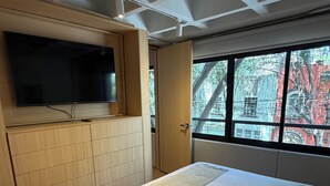 Ein Schlafzimmer mit einem Bett, einem an einer Holzkommode befestigten Fernseher und einem Fenster mit Blick auf Gebäude.