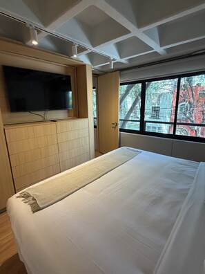 Ein Schlafzimmer mit einem Bett, einem an einer Holzkommode befestigten Fernseher und einem Fenster mit Blick auf Gebäude.