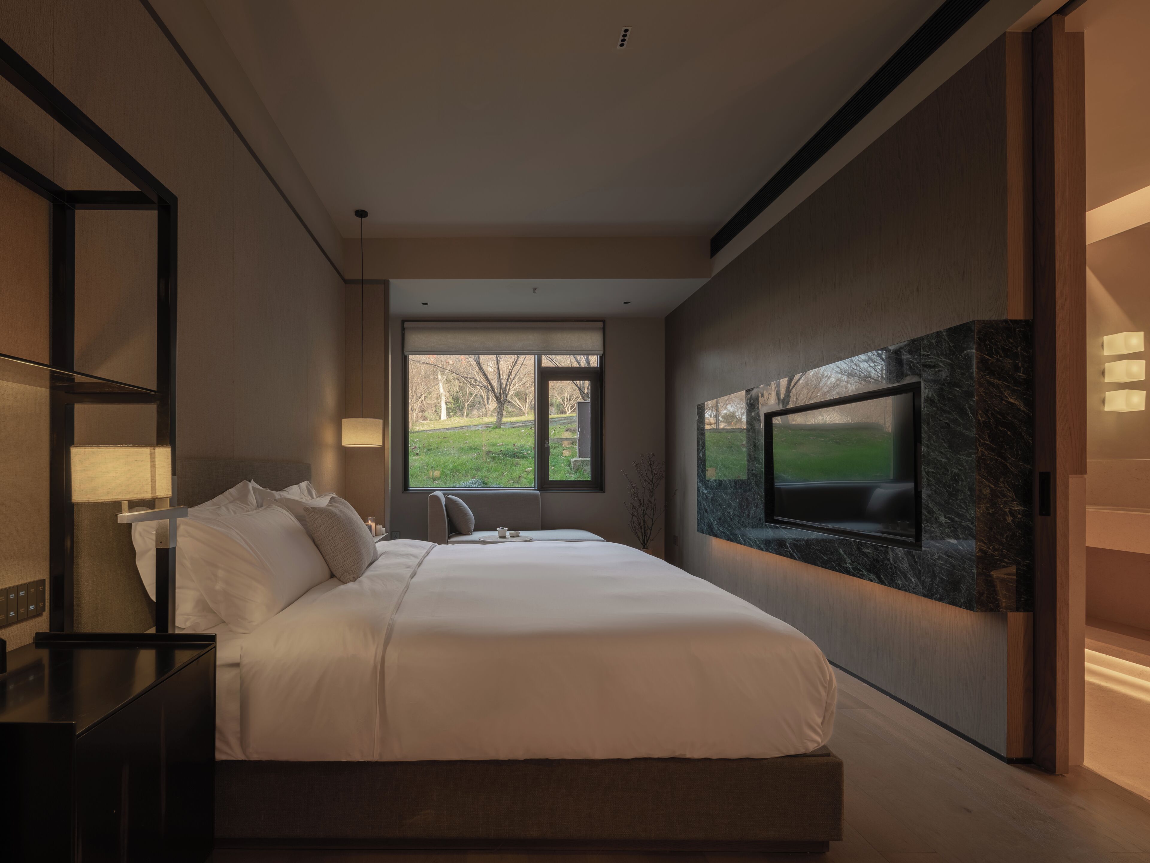 Un dormitorio moderno con una cama grande, un televisor montado en una pared de mármol y una ventana con vistas al exterior.