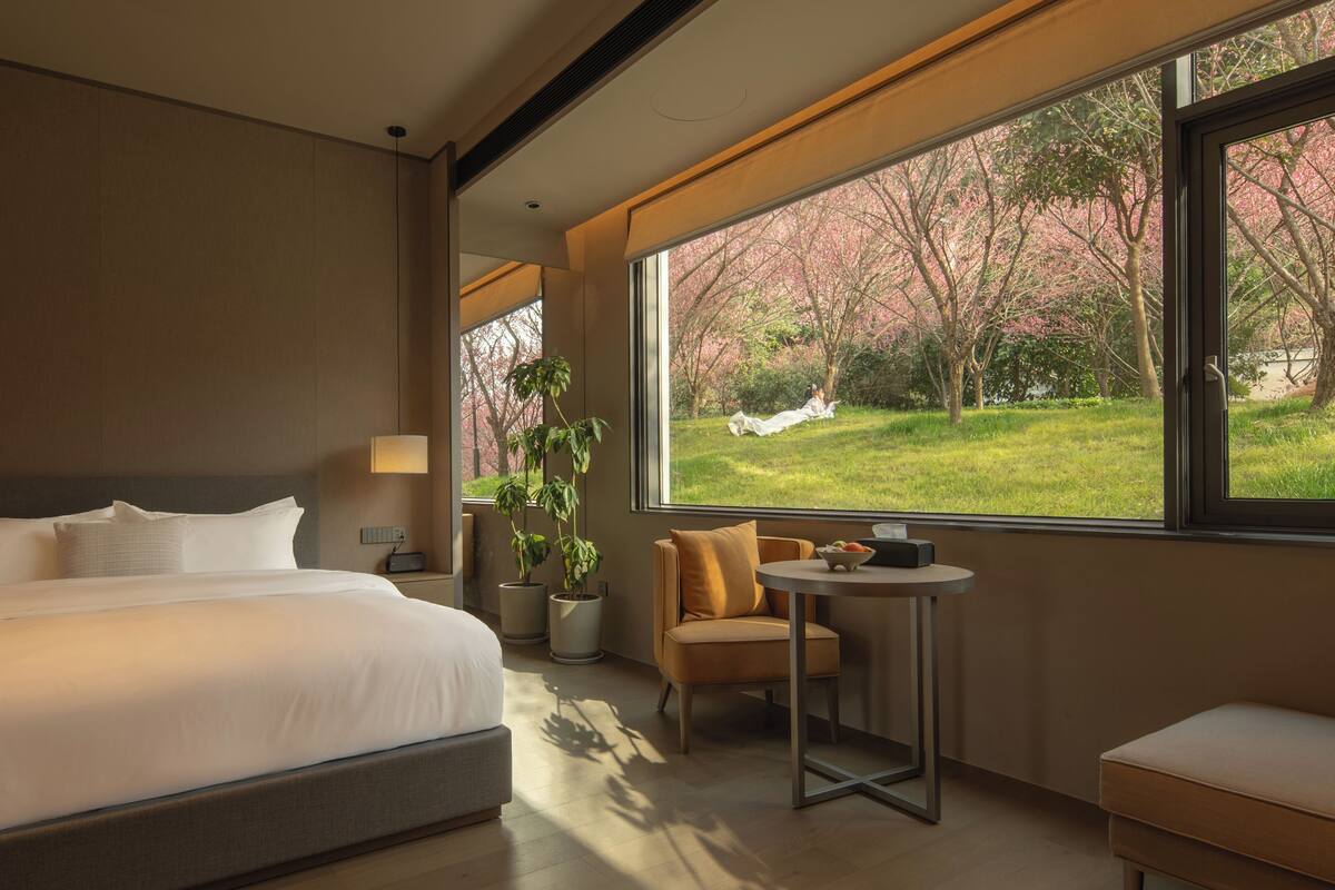 Quarto de hotel com uma cama grande, uma mesinha, uma cadeira e vista para cerejeiras.